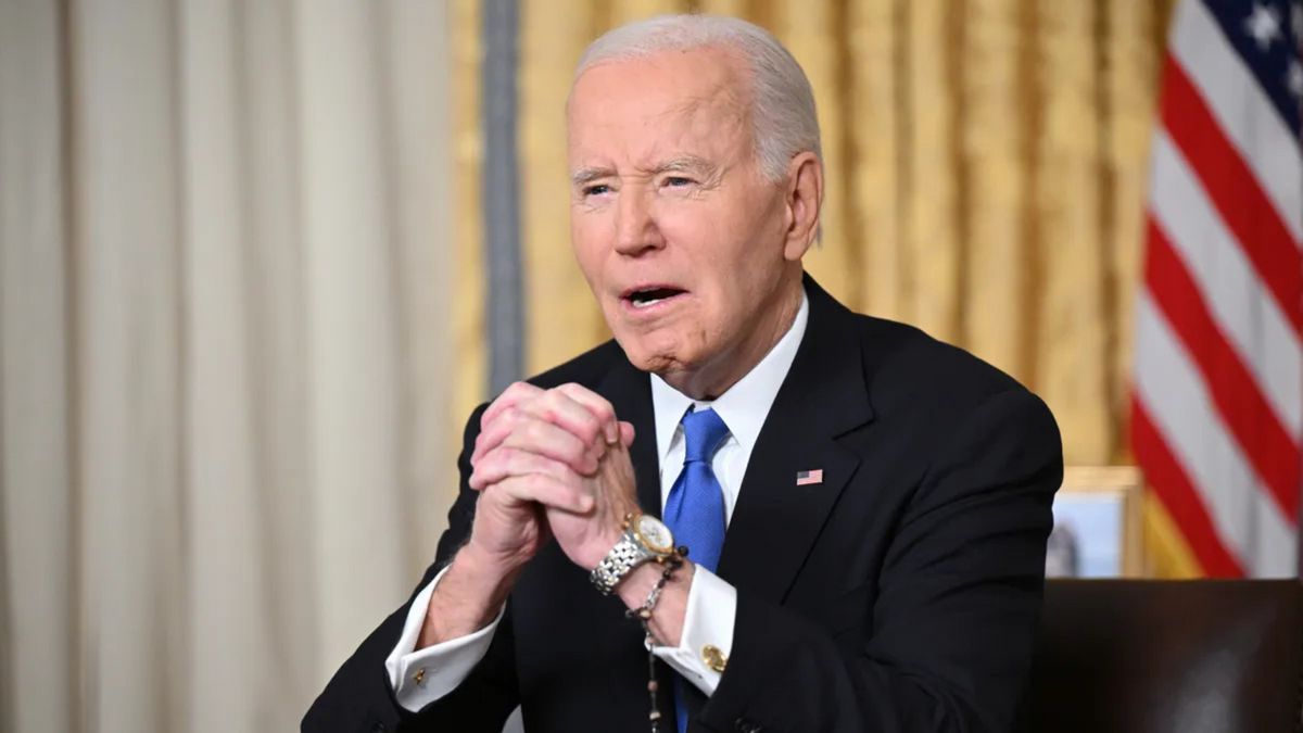 A white man wearing a dark blue suit sits at a desk with his hands clasped.