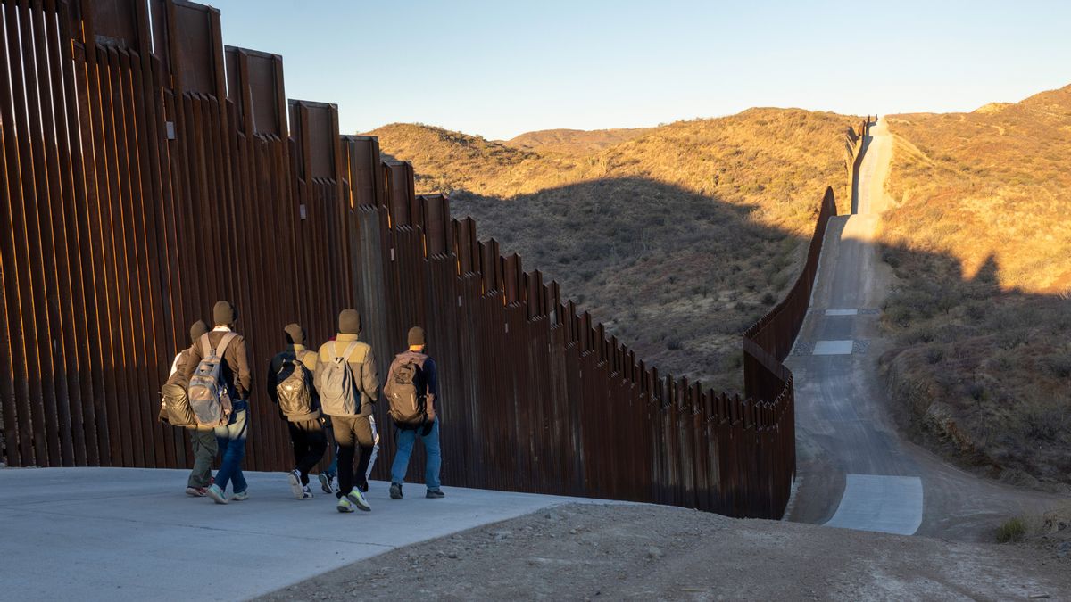 A group of people walk along a border wall.