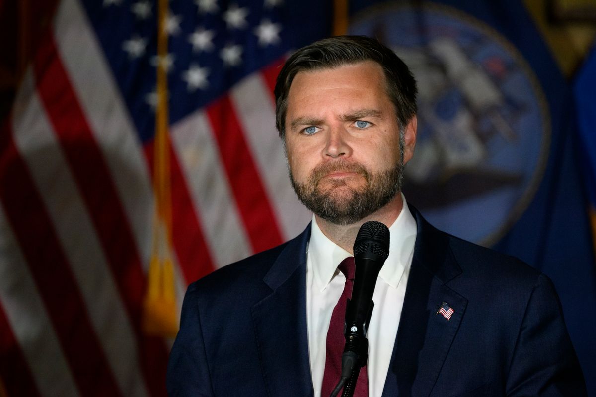 A man with blue eyes and short cropped brown hair wearing a dark blue suit and red tie with a United States flag lapel. 