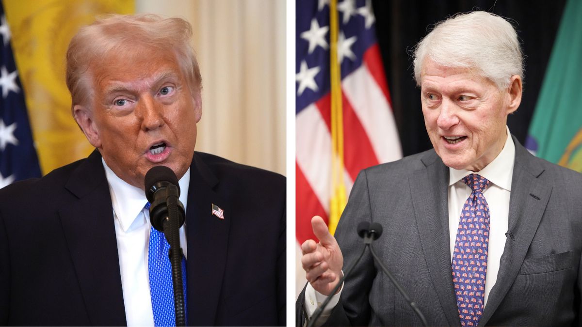 A white man wearing a dark suit is pictured next to another white man wearing a gray suit. The man on the left is speaking at a microphone, while the man on the right is looking towards the left while he also speaks into a microphone.