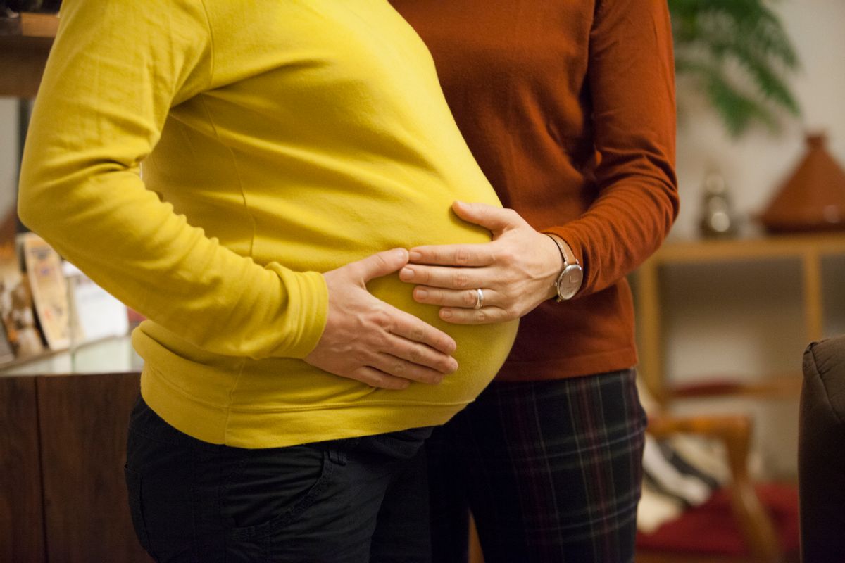A pregnant person in a yellow long-sleeved shirt stands in profile (their head is not in view). Another person in a long-sleeved rust orange shirt wearing a watch and with a ring on their ring finger places their hand on the pregnant person's belly.