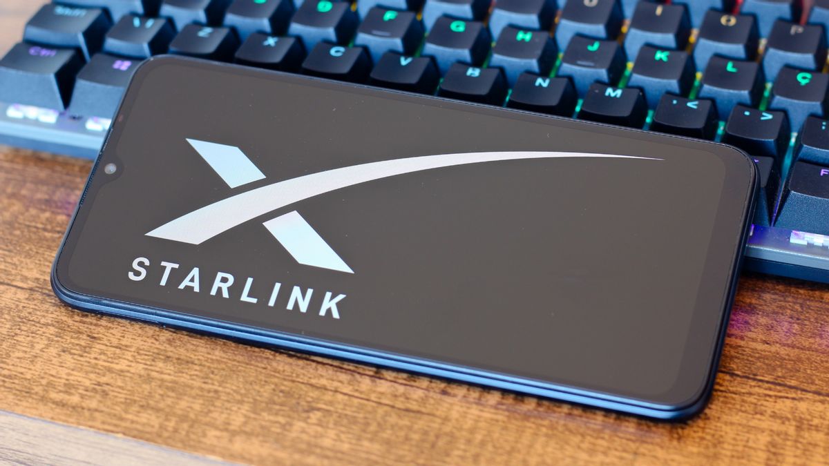 A phone displays a X logo above a white word that says STARLINK. It horizontally rests partially on a keyboard, partially on a desk.
