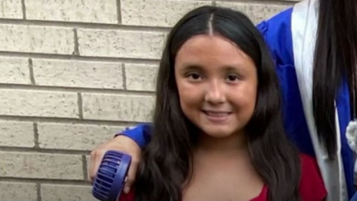A Hispanic girl smiles in front of a wall. Someone stands to her right.