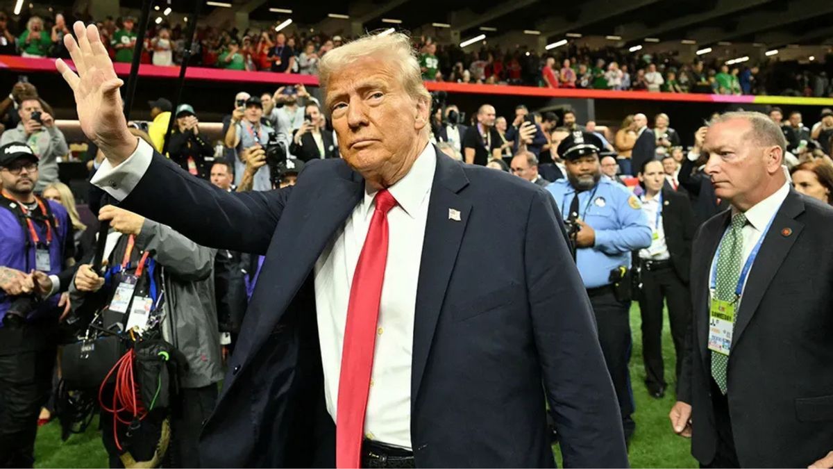 A white man wearing a dark blue suit looks toward the camera while raising his left hand in a wave. He stands on a football field; a crowd of people stand around him.