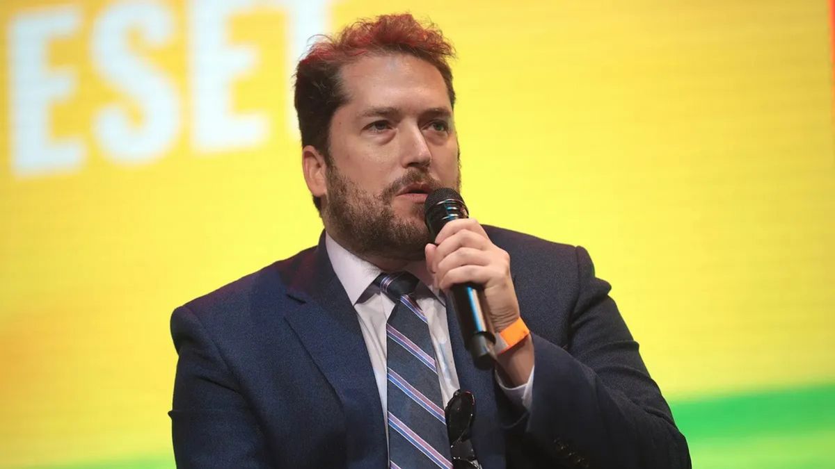A white man wearing a blue suit speaks into a microphone.