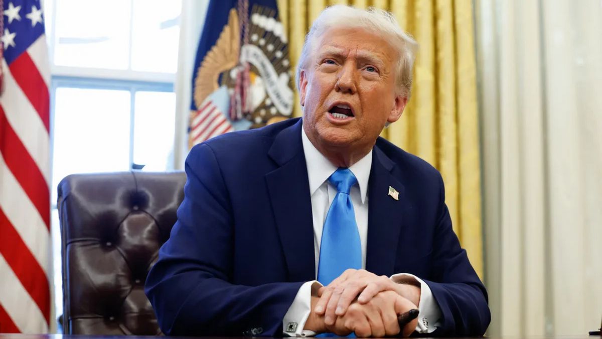 A white man wearing a blue suit sits at a desk with his right hand held over the left.