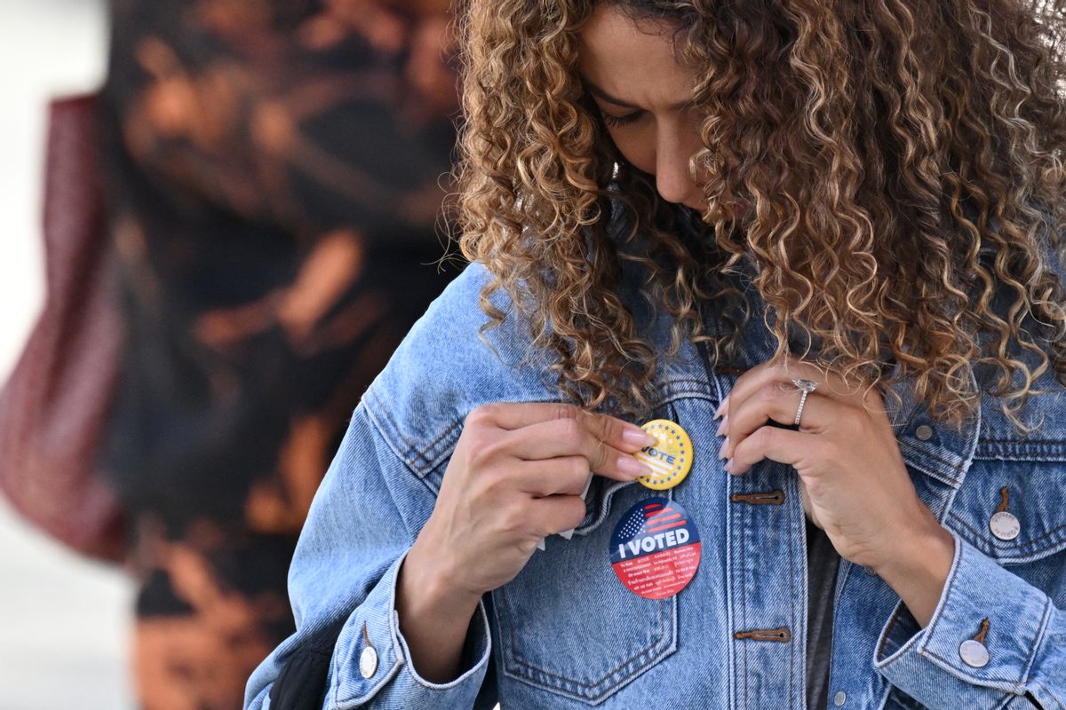 A woman with long curly brown hair with blonde highlights attaches a pin to her blue denim jacket. An "I VOTED" sticker sits below the pin. 