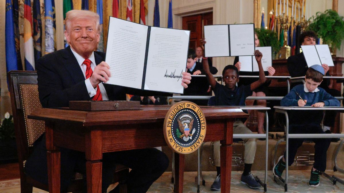 U.S. President Donald Trump holds up an executive order while sitting behind a desk with the presidential seal. Kids behind him are doing the same at their own desks.