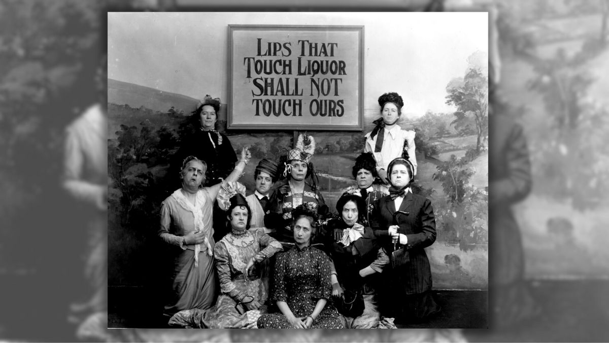 A group of people sit and stand around a sign that says "LIPS THAT TOUCH LIQUOR SHALL NOT TOUCH OURS."