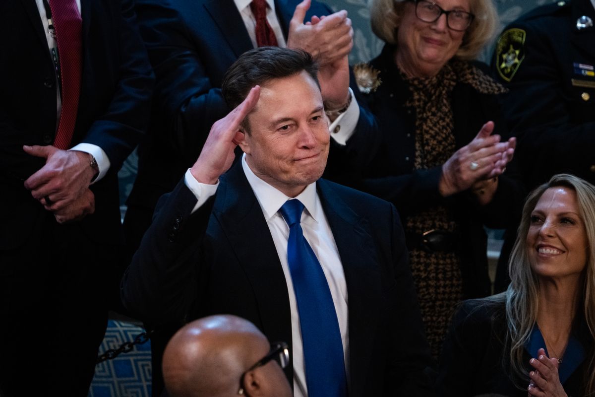A white man with short brown hair in a suit stands up and salutes; he is surrounded by seated people clapping. 