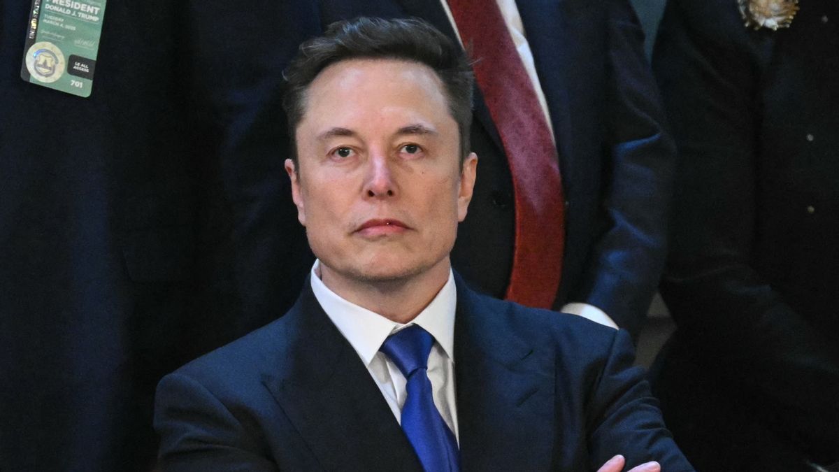 A whie man wearing a blue suit stands with his arms crossed over his chest.