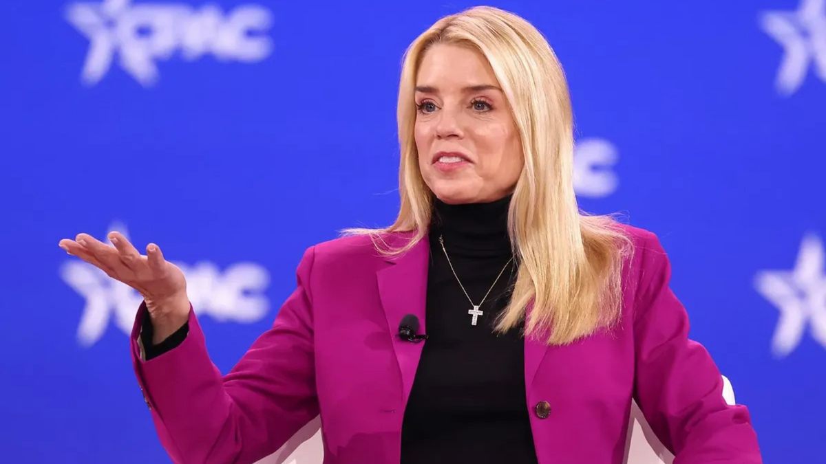 A white woman wearing a cross necklace and dark pink jacket holds her left hand out, palm up.