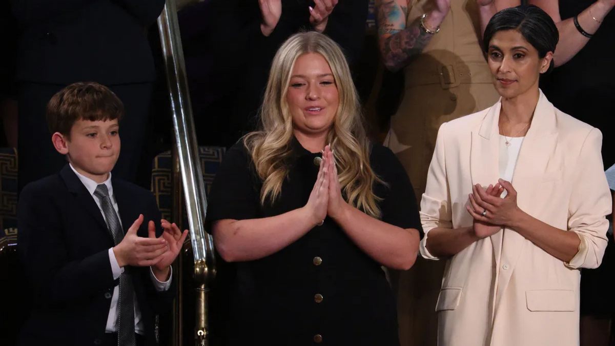 A white boy, white woman and Indian woman all stand with their hands in various states of applause. The woman in the middle has her hands pressed against each other.
