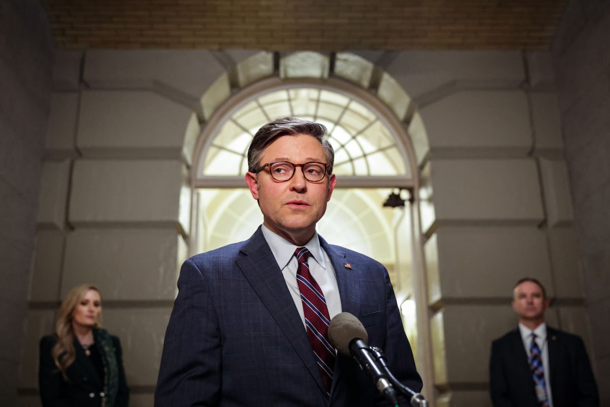 A white man with short cropped brown hair wearing a dark suit standing in front of a microphone. 