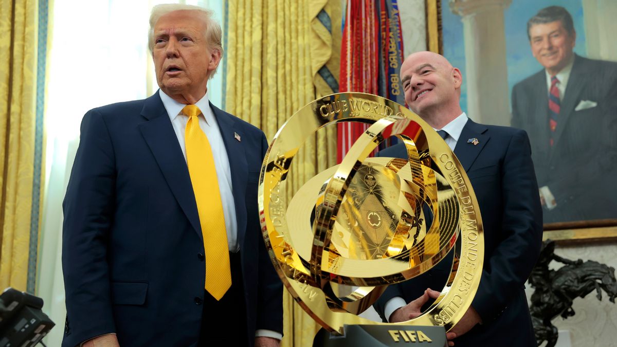 A white man wearing a blue suit stands next to another white man wearing a blue suit. A large gold award that says FIFA in gold letters at the bottom stands between them. A portrait of a white man wearing a black suit is seen on the wall.