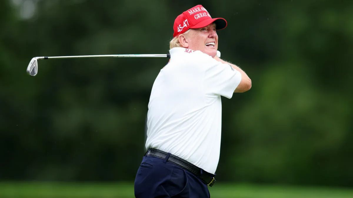A white man wearing a white shirt and red hat that says MAKE AMERICA GREAT AGAIN in white letters smiles while holding a golf club.
