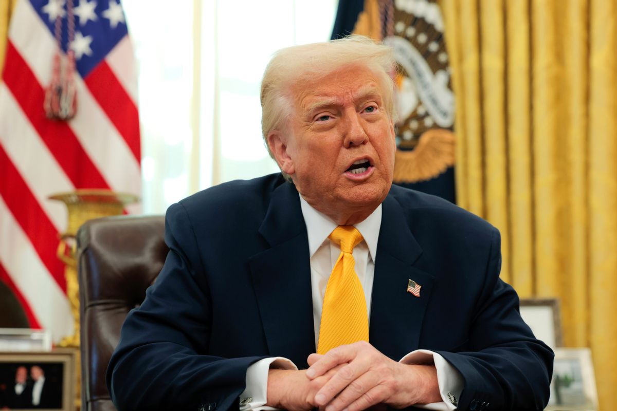 U.S. President Donald Trump, a white man with a dark tan and pale blonde hair wearing a dark suit and yellow tie, speaks with his hands crossed while seated in front of a U.S. flag and the U.S. seal. 