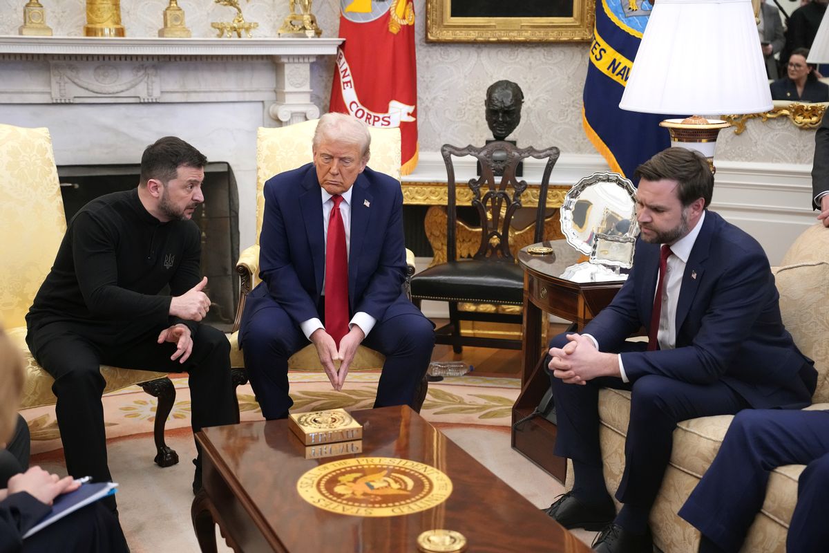 Three white men sit in a room. The one to the left is wearing a black long-sleeved shirt and pants, while the other two men are wearing dark blue suits.
