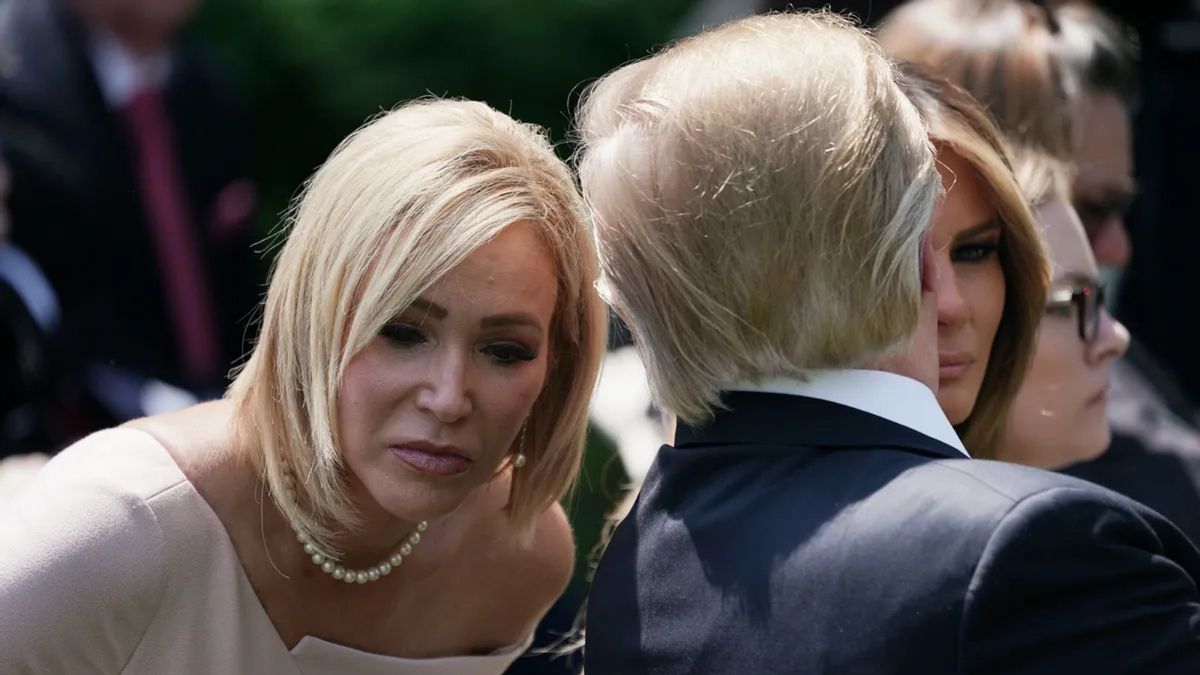 A white woman with a blonde bob wearing a white dress leans forward. A blonde man, pictured from behind, speaks into her ear. 