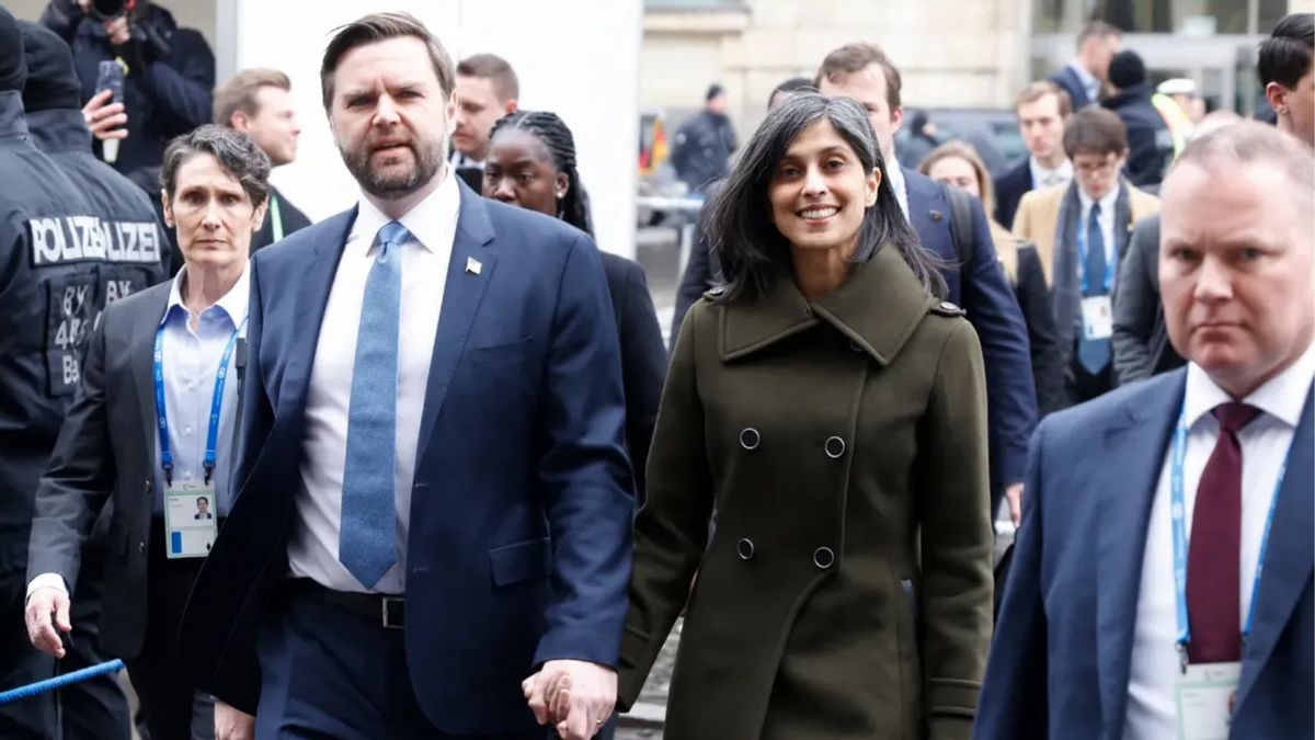 A white man wearing a blue suit walks holding hands with an Indian woman wearing a pea green coat and smiling at the camera. They are surrounded by people walking around them.