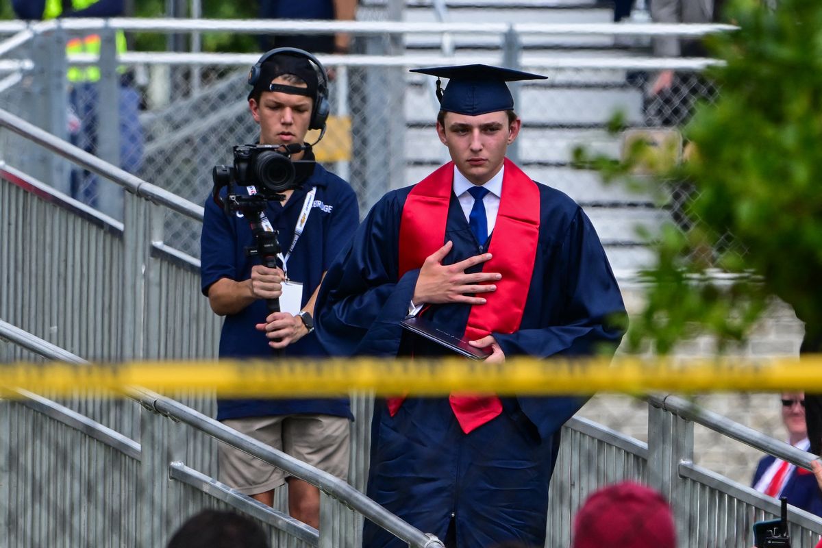 A white teenager in a graduation cap and gown with a red stole stands near a railing. 