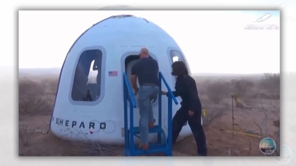 A white man stands in front of a rocket capsule's hatch. A person stands behind him.