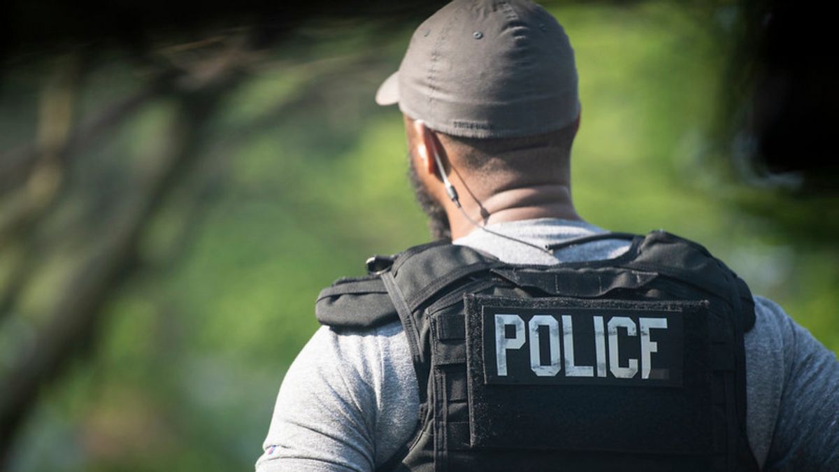A person wears a vest that says POLICE on their back. They stand outside.