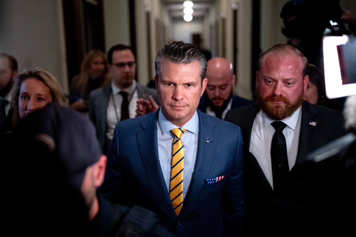 A white man with greying hair in a blue suit and yellow striped tie walks down a hallway, surrounded by other people in suits. 