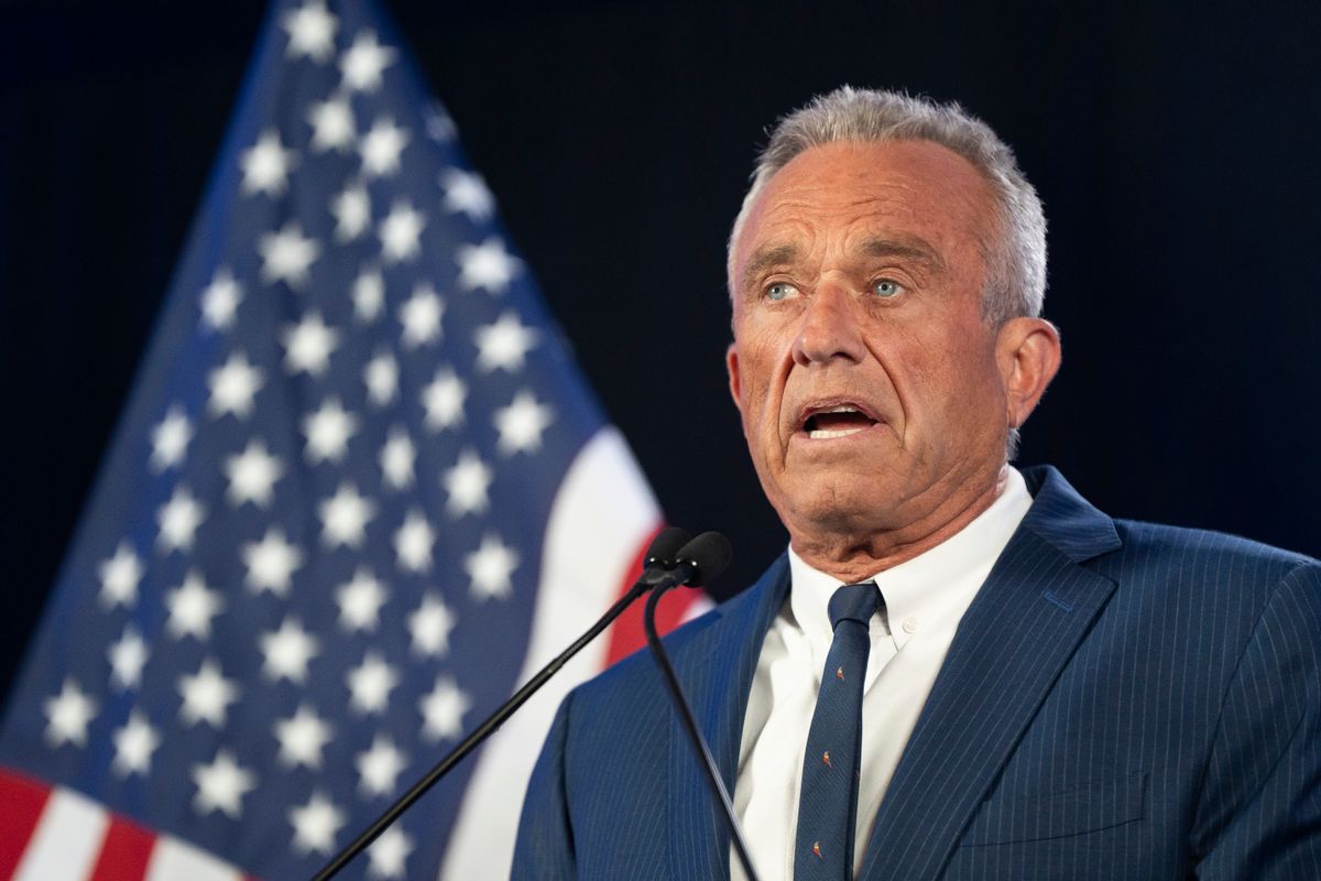 A white tan man with short grey and white hair in his 70s wearing a suit speaks in front of a microphone. 