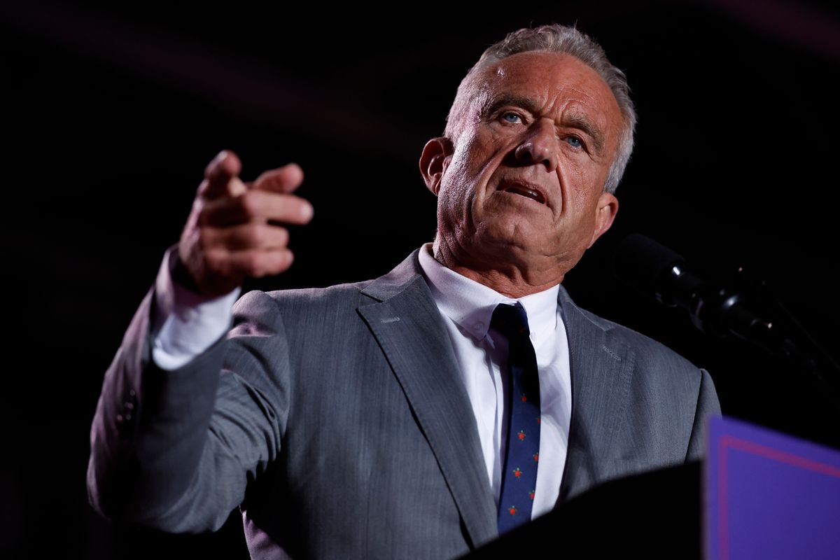 A white man with greying hair and a dark tan points at the camera. He is wearing a grey suit with a dark tie and standing behind a podium. 