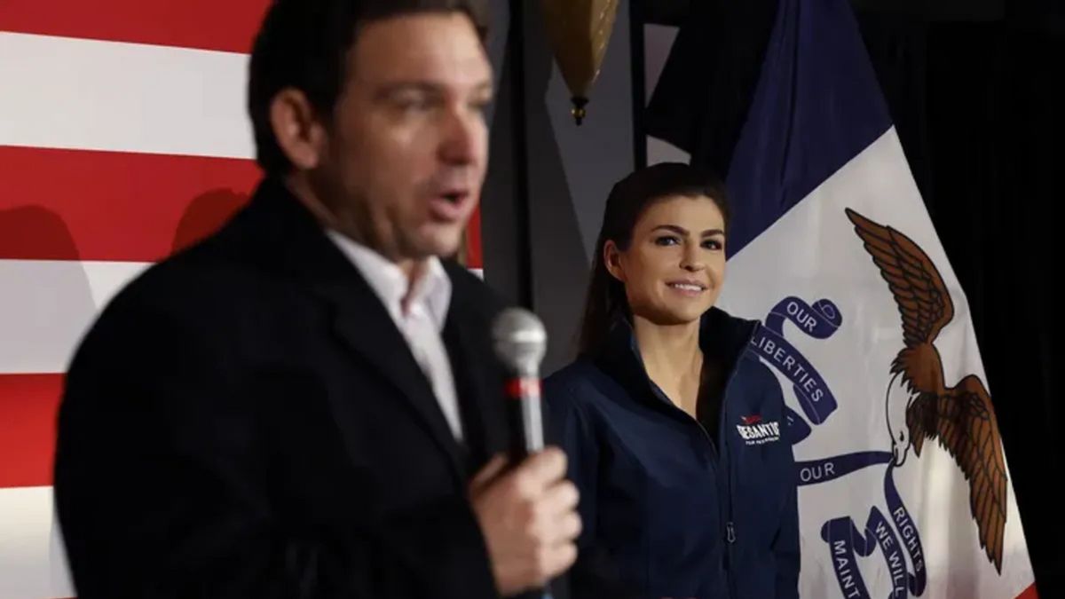 A white woman wearing a blue jacket looks at a white man wearing a black suit speaking into a microphone. The camera is focused on the woman.