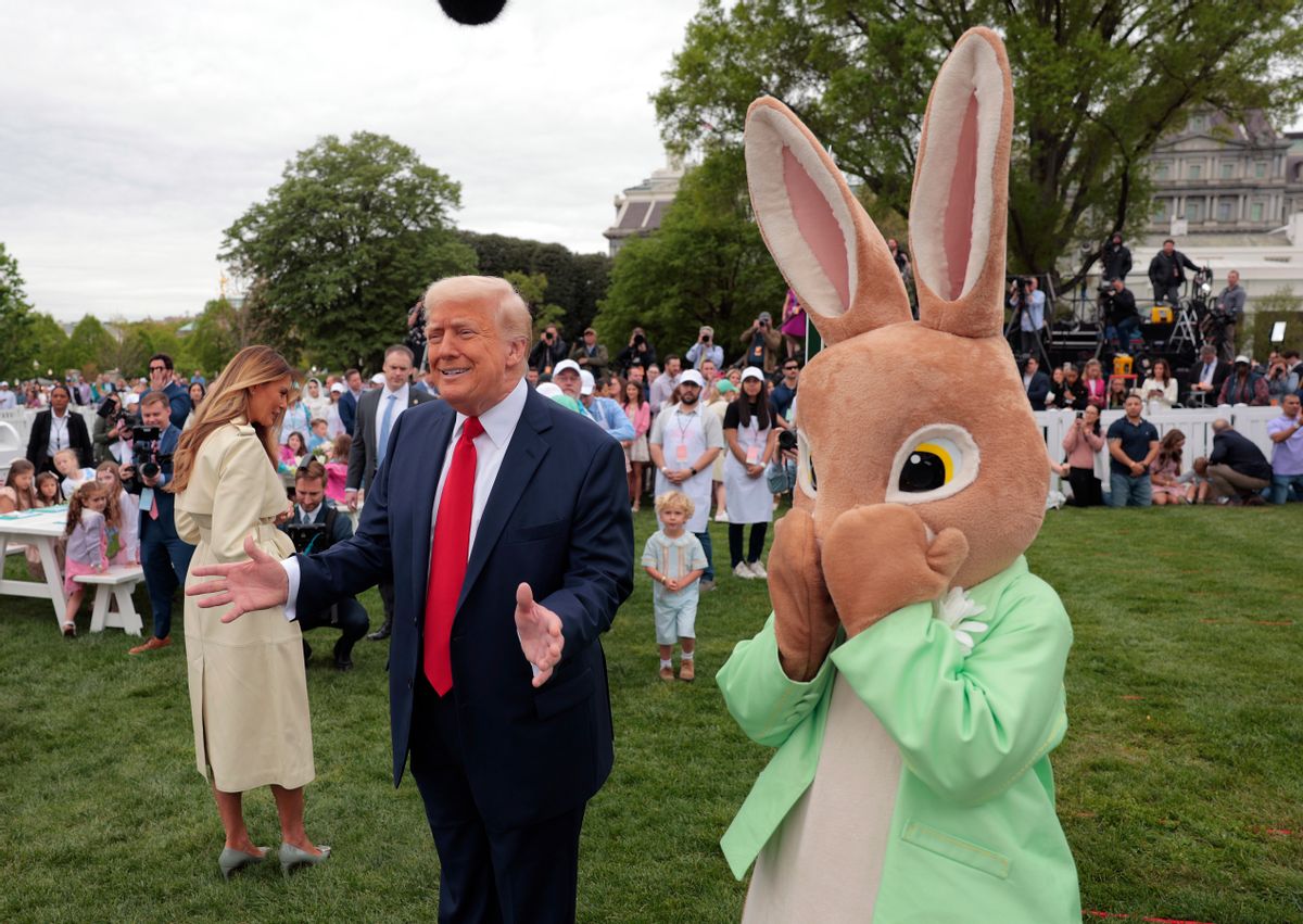 A white man holds his arms open next to a large Easter bunny. A white woman stands with her back to the camera. A large group of people is seen behind them.