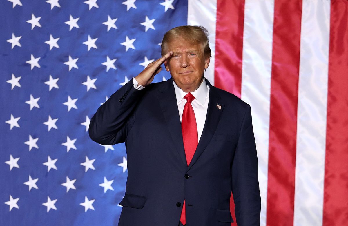 A white man wearing a dark suit salutes in front of a large red, white and blue flag.