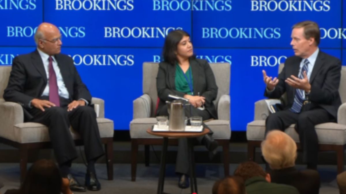 Three people sit on chairs in front of screens that say "BROOKINGS." Two of the people on the left look at the person sitting on the very right.
