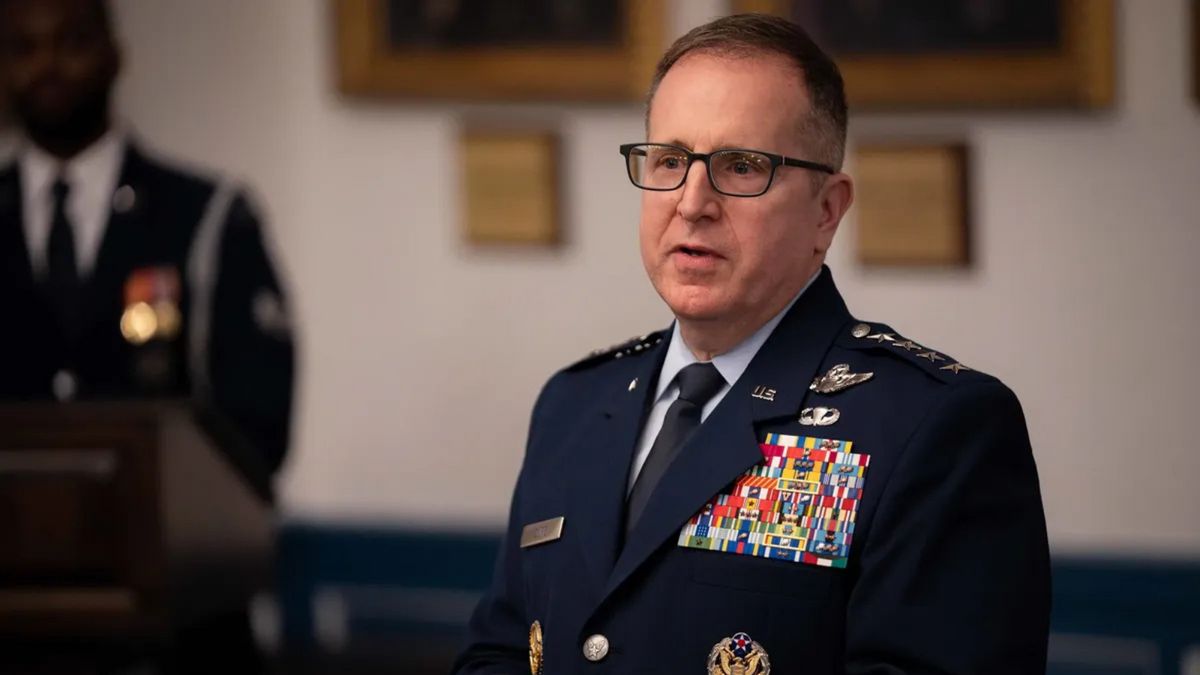 A white man wearing a blue uniform stands looking to the left. You can see another person standing in the background out of focus.
