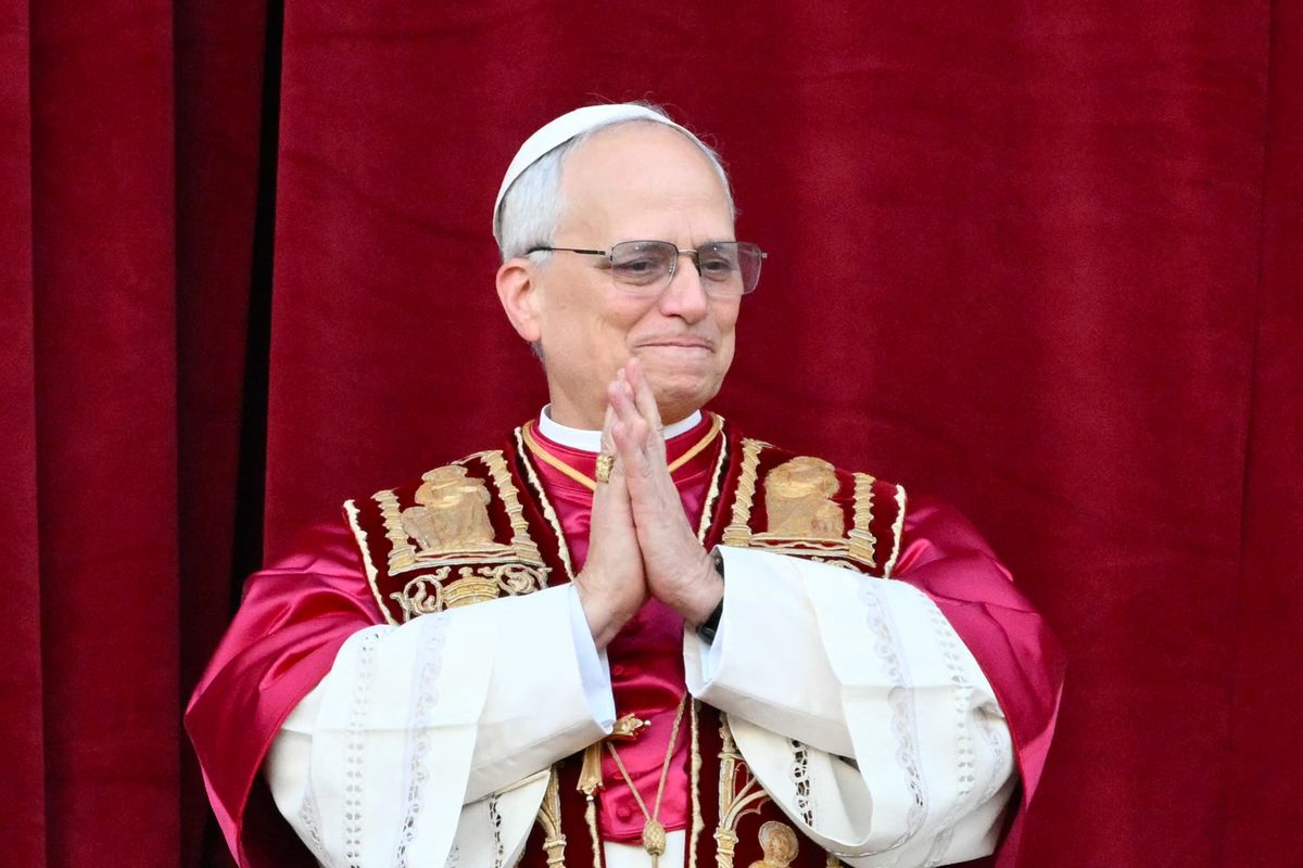 A white man with short white receding hair wearing glasses clasps his hands together. He is wearing a traditional red and gold papal robe and a small white skullcap. 