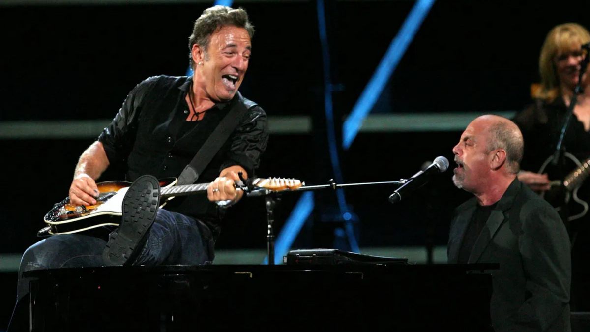 Two white men perform on stage. One holds a guitar and sits on a piano looking to his right at the person singing sitting on its bench.