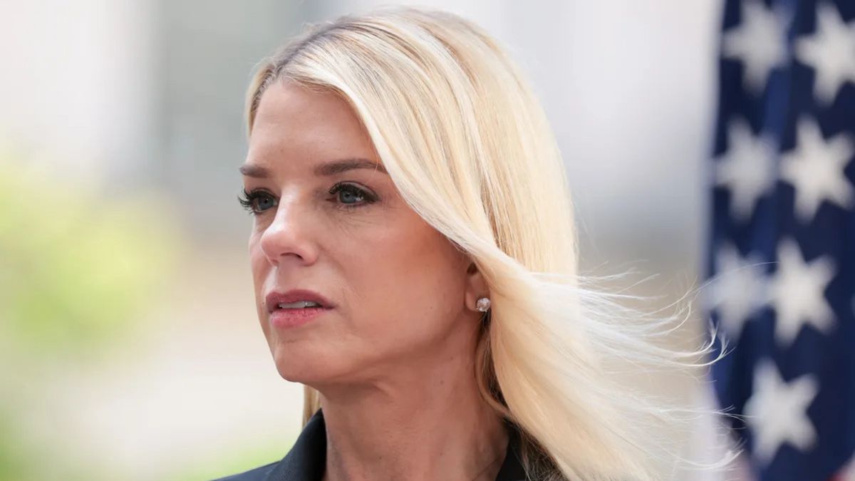 A white woman looks forward. A red, white and blue flag can be seen in the background.