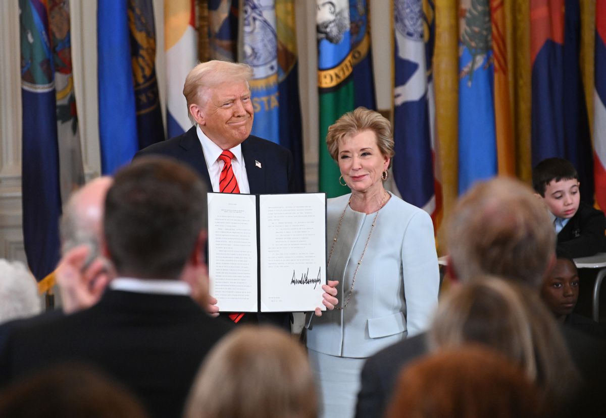 U.S. President Donald Trump, a white man with blonde hair in a suit, stands next to a white woman with short blonde hair in a light blue dress. He is holding up a signed document.