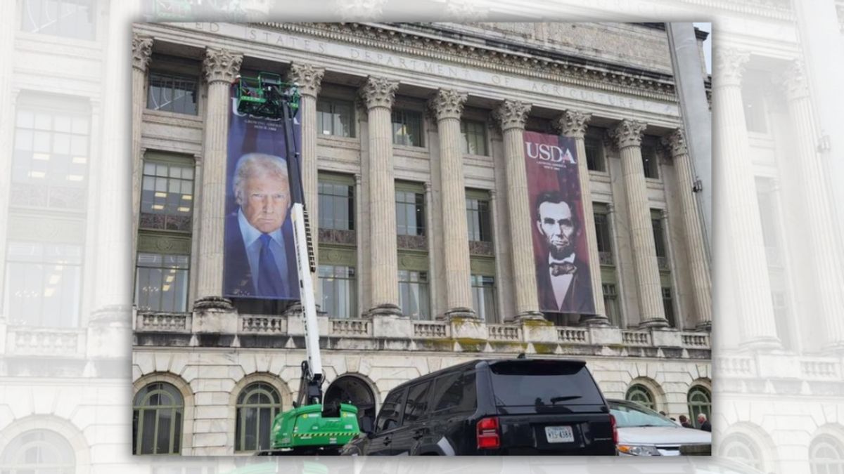 A banner of a white man is shown being placed on a white building. It's next to a banner of another white man to his right. Two cars can be seen in front of the building.