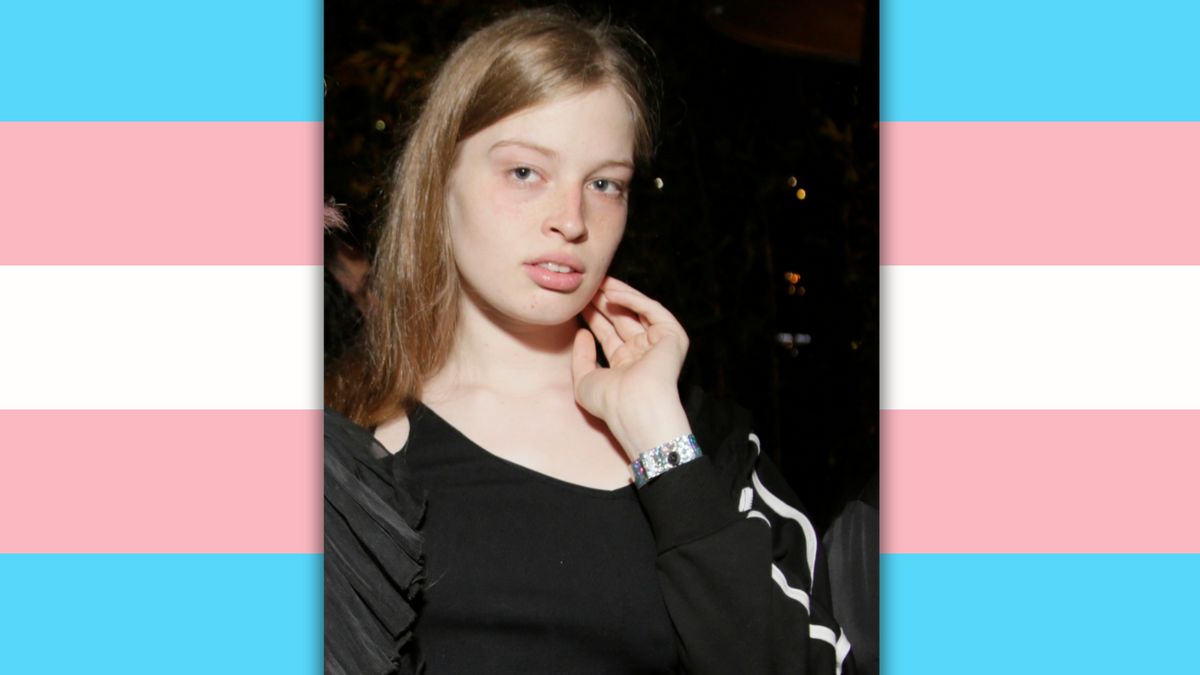 A white woman with long blonde hair poses for the camera. Behind her is a pink, blue and white striped trans pride flag. 