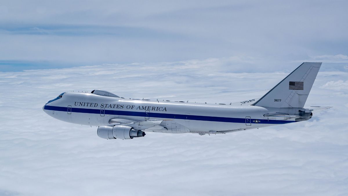 A U.S. Air Force E-4B "Nightwatch" aircraft, known as the "Doomsday Plane," flies above a blanket of clouds. The aircraft is painted white with a blue stripe and the words "United States of America" along the fuselage. The American flag and tail number 31677 are visible on the vertical stabilizer.