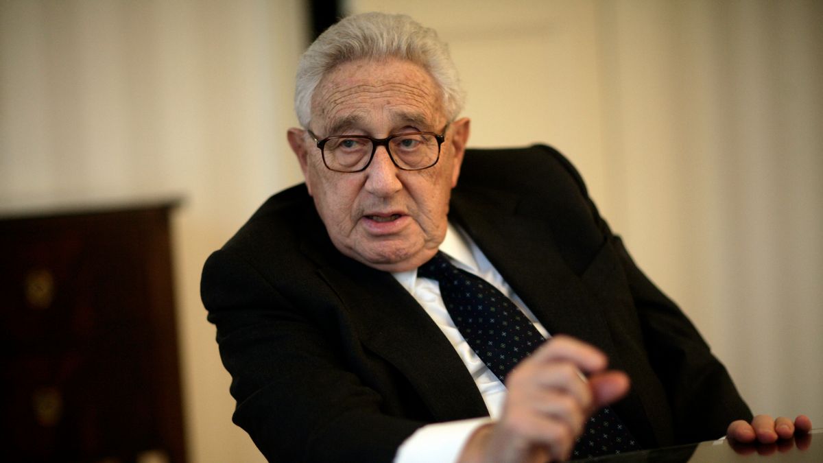 Henry Kissinger, wearing a suit, blue tie, and glasses, speaks during an interview in Washington, D.C. He is seated in a chair, gesturing with one hand, with a neutral backdrop behind him.