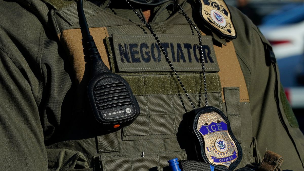 Close-up of a U.S. Immigration and Customs Enforcement (ICE) officer's tactical vest. The vest features a large patch labeled “NEGOTIATOR,” a Motorola radio microphone clipped to the front, and a metallic ICE badge hanging from a beaded chain. The officer is wearing a green uniform.