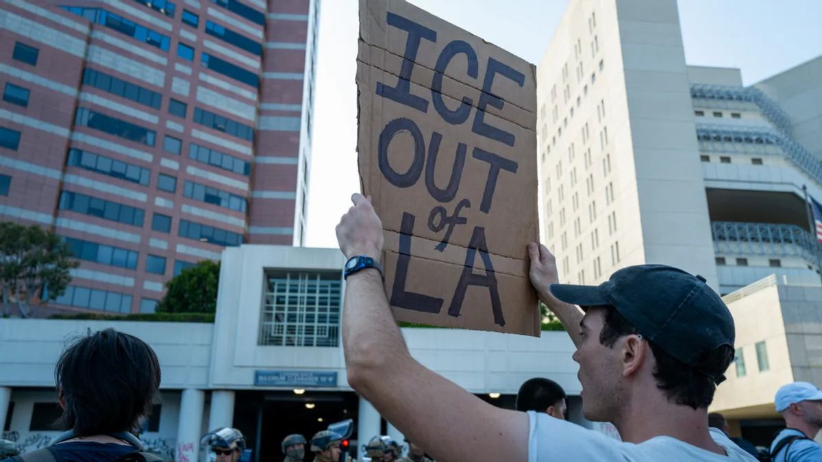 A person holds a sign that says "ICE OUT of LA." Other people can be seen around them, as well as soldiers faintly towards the bottom of the image.