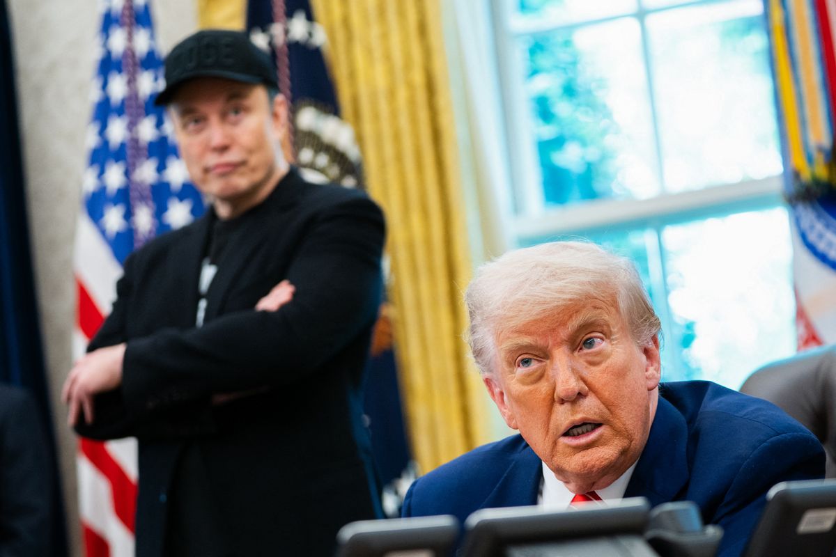 A white man wearing a blue suit is sitting down while speaking. Behind him, a white man wearing all black can be seen slightly blurred in the background.