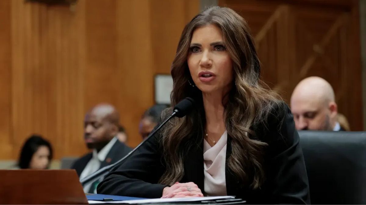 A white woman speaks into a microphone. Several people can be seen sitting behind her.