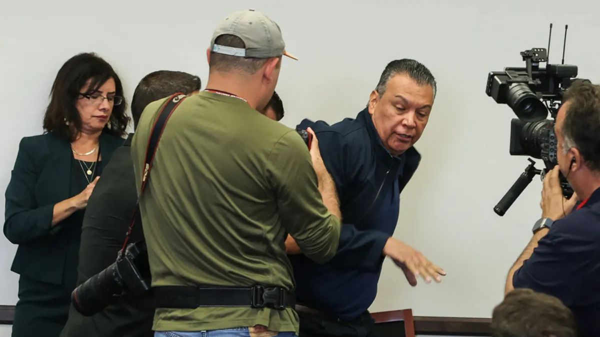 Several people surround a Latino wearing a dark blue jacket. Two of the people appear to hold cameras.