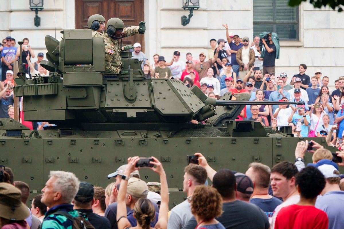A large crowd surrounds a tank with two soldiers standing on it waving to the crowd.