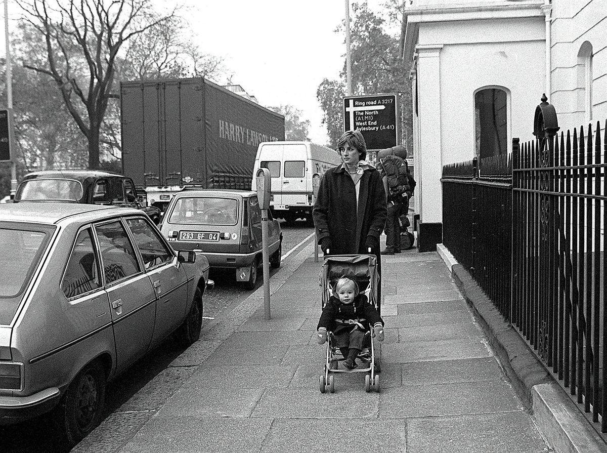 In this black and white image, a young woman pushes a stroller with a baby inside along a sidewalk. 